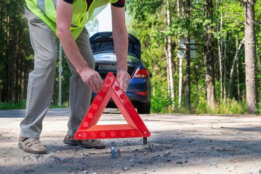 An adult retiree places a portable, reflective red triangular warning triangle on the side of a rural road near his car. Selective focus.