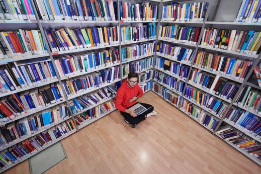 a young student with glasses sits in the library, reads a book and makes notes with a latpop