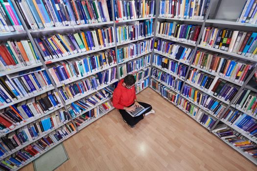 a young student with glasses sits in the library, reads a book and makes notes with a latpop