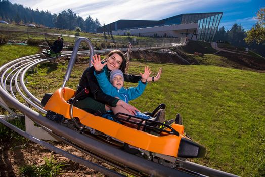 Excited young mother and son driving on alpine coaster while enjoying beautiful sunny day in the nature