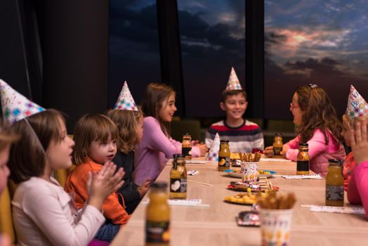 young happy boy and group of his friends having birthday party with a night sky through the windows in the background