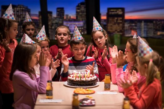 young happy boy and group of his friends having birthday party with a night city through the windows in the background