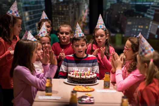 young happy boy and group of his friends having birthday party with a night city through the windows in the background