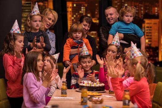 young happy boy and group of his friends having birthday party with a night city through the windows in the background