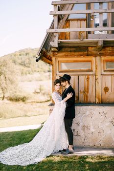 Groom hugs bride in a white lace dress near the wooden house. High quality photo