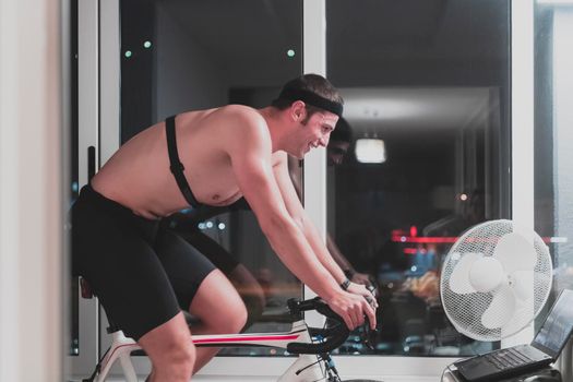 Man cycling on the machine trainer he is exercising in the home at night. Playing online bike racing game during coronavirus covid19 lockdown. New normal concept.