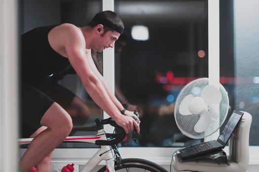 Man cycling on the machine trainer he is exercising in the home at night. Playing online bike racing game during coronavirus covid19 lockdown. New normal concept.