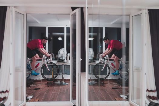 Man cycling on the machine trainer he is exercising in the home at night. Playing online bike racing game during coronavirus covid19 lockdown. New normal concept.