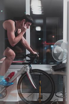 Man cycling on the machine trainer he is exercising in the home at night. Playing online bike racing game during coronavirus covid19 lockdown. New normal concept.