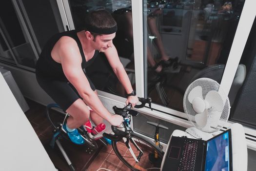 Man cycling on the machine trainer he is exercising in the home at night playing online bike racing game during coronavirus covid19 lockdown new normal