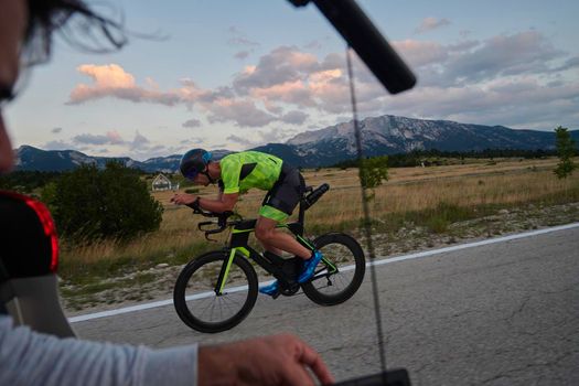 cinematographer with professional cinema camera taking sport action shot  from moving car of triathlon athlete who riding bike on low traffic country road