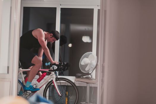 Man cycling on the machine trainer he is exercising in the home at night playing online bike racing game during coronavirus covid19 lockdown new normal