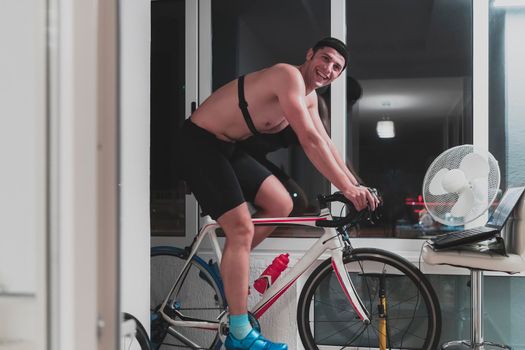 Man cycling on the machine trainer he is exercising in the home at night. Playing online bike racing game during coronavirus covid19 lockdown. New normal concept.