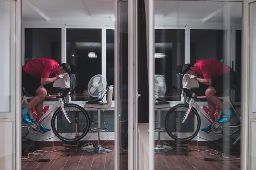 Man cycling on the machine trainer he is exercising in the home at night. Playing online bike racing game during coronavirus covid19 lockdown. New normal concept.