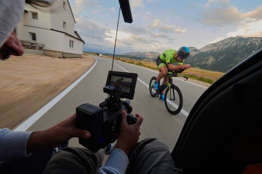 cinematographer with professional cinema camera taking sport action shot  from moving car of triathlon athlete who riding bike on low traffic country road