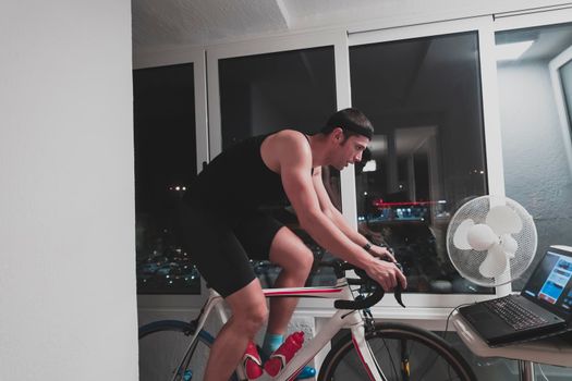 Man cycling on the machine trainer he is exercising in the home at night. Playing online bike racing game during coronavirus covid19 lockdown. New normal concept.