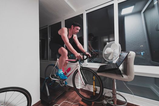 Man cycling on the machine trainer he is exercising in the home at night. Playing online bike racing game during coronavirus covid19 lockdown. New normal concept.