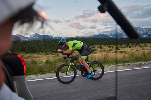 cinematographer with professional cinema camera taking sport action shot  from moving car of triathlon athlete who riding bike on low traffic country road