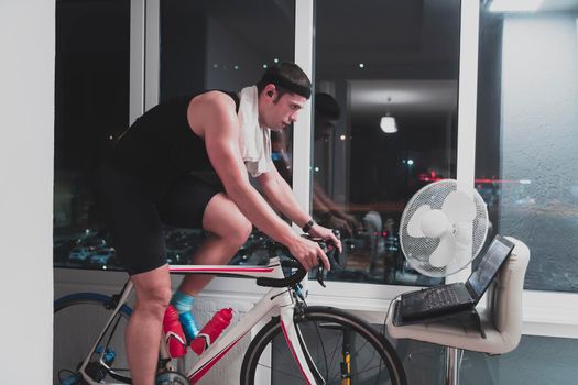 Man cycling on the machine trainer he is exercising in the home at night. Playing online bike racing game during coronavirus covid19 lockdown. New normal concept.