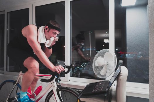 Man cycling on the machine trainer he is exercising in the home at night. Playing online bike racing game during coronavirus covid19 lockdown. New normal concept.