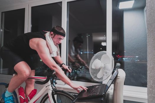 Man cycling on the machine trainer he is exercising in the home at night. Playing online bike racing game during coronavirus covid19 lockdown. New normal concept.