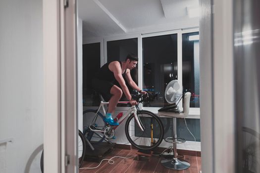 Man cycling on the machine trainer he is exercising in the home at night playing online bike racing game during coronavirus covid19 lockdown new normal