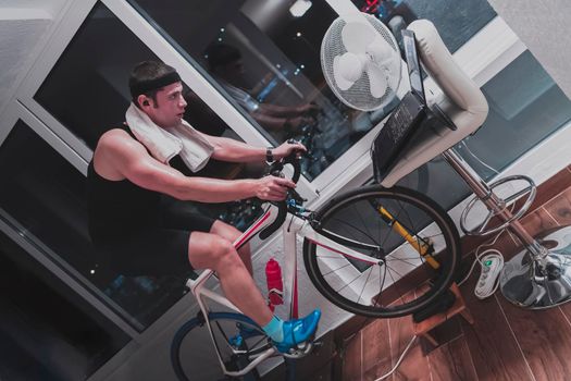 Man cycling on the machine trainer he is exercising in the home at night. Playing online bike racing game during coronavirus covid19 lockdown. New normal concept.