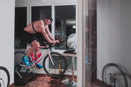 Man cycling on the machine trainer he is exercising in the home at night. Playing online bike racing game during coronavirus covid19 lockdown. New normal concept.