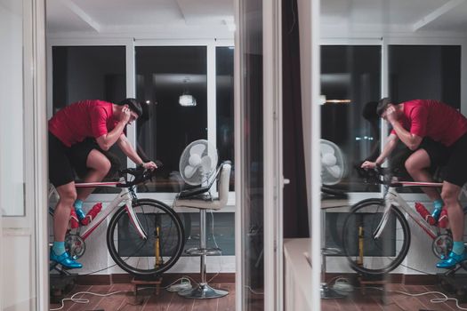 Man cycling on the machine trainer he is exercising in the home at night. Playing online bike racing game during coronavirus covid19 lockdown. New normal concept.