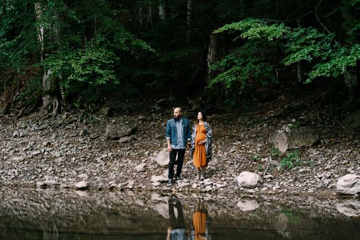 Pregnant woman with man stands on a rocky slope by a lake surrounded by trees. High quality photo