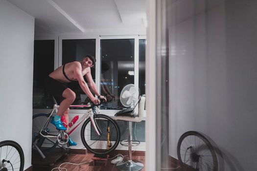 Man cycling on the machine trainer he is exercising in the home at night. Playing online bike racing game during coronavirus covid19 lockdown. New normal concept.