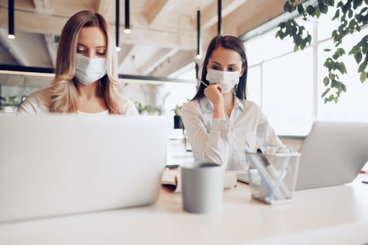 Two female colleagues working in office together wearing medical masks, coronavirus prevention