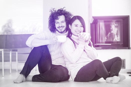 Young romantic multiethnic couple sitting on the floor in front of fireplace at home, talking and drinking coffee at cold winter day