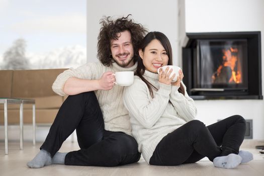 Young romantic multiethnic couple sitting on the floor in front of fireplace at home, talking and drinking coffee at cold winter day