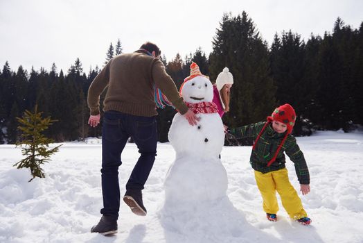 happy young  family playing in fresh snow and building snowman at beautiful sunny winter day outdoor in nature