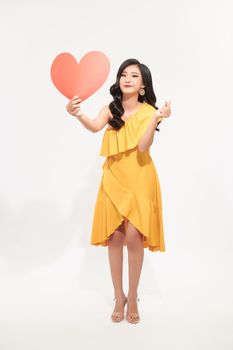 Portrait of a happy young woman dressed in yellow dress holding paper heart above her head isolated over white background