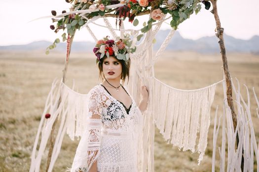 Bride in a braided dress and a wreath stands near the wedding arch in the field. High quality photo