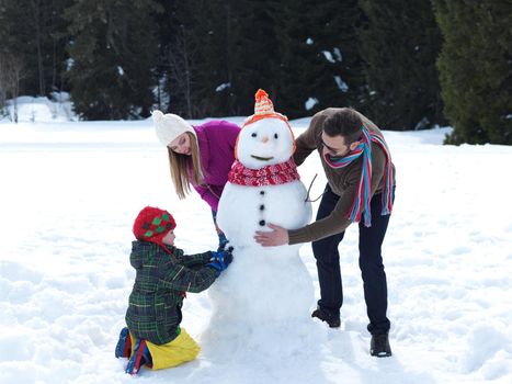 happy young  family playing in fresh snow and making snowman at beautiful sunny winter day outdoor in nature with forest in background