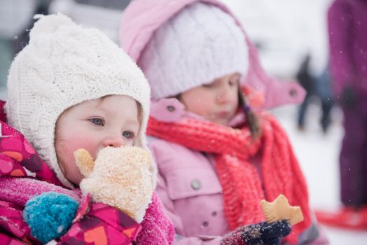 portrait of two cute little girls sitting together on sledges outdoors at snowy winter day, eating tasty cookies on break