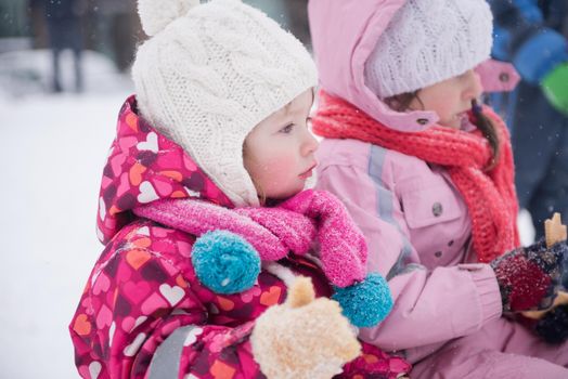 portrait of two cute little girls sitting together on sledges outdoors at snowy winter day, eating tasty cookies on break