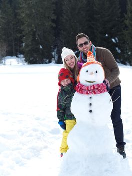 happy young  family playing in fresh snow and making snowman at beautiful sunny winter day outdoor in nature with forest in background