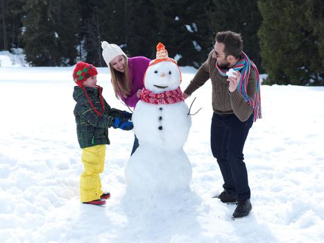 happy young  family playing in fresh snow and making snowman at beautiful sunny winter day outdoor in nature with forest in background