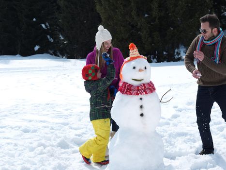 happy young  family playing in fresh snow and making snowman at beautiful sunny winter day outdoor in nature with forest in background