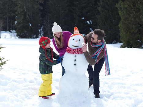 happy young  family playing in fresh snow and making snowman at beautiful sunny winter day outdoor in nature with forest in background