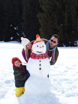 happy young  family playing in fresh snow and making snowman at beautiful sunny winter day outdoor in nature with forest in background