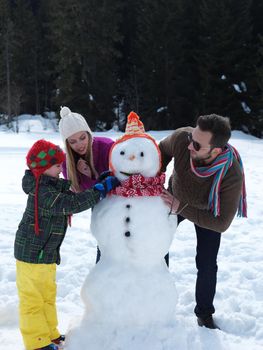 happy young  family playing in fresh snow and making snowman at beautiful sunny winter day outdoor in nature with forest in background