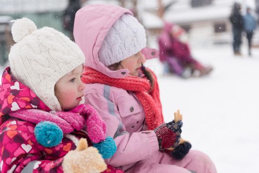 portrait of two cute little grils sitting together on sledges outdoors at snowy winter day, eating tasty cookies on break