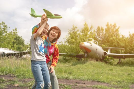 Mother and daughter child playing with toy plane at the field together with real airplane on background and smiling. Happy family moments at the nature
