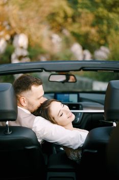 Groom hugs bride who has laid her head on his shoulder in a convertible. High quality photo