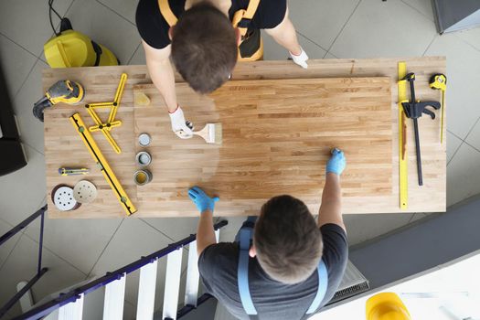 Top view of coworkers covering wooden plank with shiny polish creative process in workshop. Male carpenters finish masterpiece in studio. Design concept